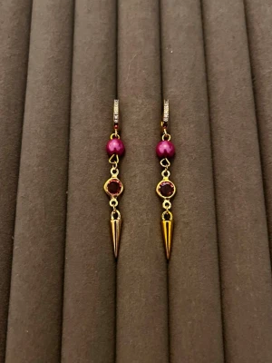 Guldörhängen med lila pärlor och stenar - Säljer ett par unika örhängen i guldfärgad metall med lila pärlor, matchande lila stenar och spetsiga hängen. Örhängena har små gnistrande stenar på bygeln och en cool, edgy design som sticker ut. Perfekta för dig som gillar detaljer och färg.