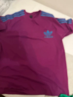 Lila Adidas t-shirt med blå detaljer - Säljer en lila Adidas t-shirt med klassiska blå ränder på axlarna och blå logga på bröstet. T-shirten har rund hals och är kortärmad, perfekt för en sportig och avslappnad stil.