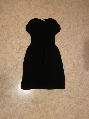 Svart kortärmad klänning - En enkel svart klänning med rund halsringning och korta ärmar. Klänningen har en markerad midja och lätt utställd kjol, vilket ger en klassisk och stilren siluett. Perfekt för dig som gillar minimalistisk stil.