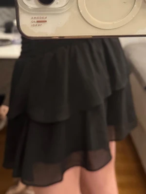 Svart volangkjol med resårmidja - Säljer en svart kortkjol med dubbla volanger och bred smockad resårmidja. Kjolen har ett lätt och flödande tyg som ger en luftig känsla. Perfekt för dig som gillar en enkel men ändå trendig look. Kan även användas som topp. Pris diskuterbart vid snabb affär💕