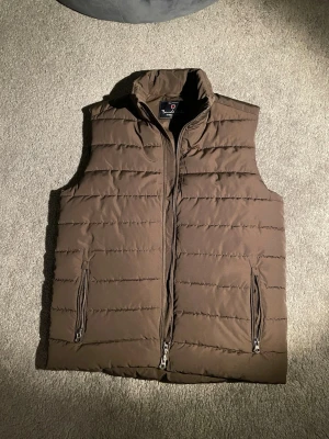 Brun dunväst med dragkedjor - Snygg brun dunväst från Bondelid med horisontella sömmar och två praktiska fickor med dragkedja. Västen har hög krage och stängs med dragkedja framtill. Perfekt för lager-på-lager och enkel att matcha med olika outfits.