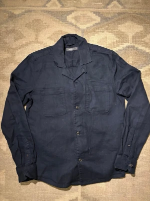 Mörkblå overshirt med fickor - Stilren mörkblå overshirt med två stora bröstfickor och knäppning framtill. Skjortan har klassisk krage och långa ärmar med manschettknappar. Perfekt att bära över en t-shirt för en avslappnad look. Materialet ser ut att vara bomull eller en bomullsblandning.