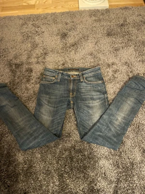 Blå slim fit jeans från Nudie Jeans - Säljer ett par blå slim fit jeans från Nudie Jeans med klassisk femficksmodell och snygga slitningar. Jeansen har normal midja, smala ben och är gjorda i mjuk denim. Perfekta för dig som gillar en avslappnad men ändå stilren look.