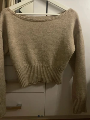 Beige croppad stickad tröja - Trendig beige croppad stickad tröja med bred ribbad mudd i midjan och vid ärmsluten. Tröjan har en bred, rundad halsringning och långa ärmar. Perfekt att styla med höga jeans eller kjol.