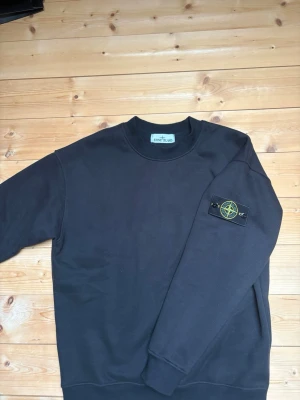 Svart sweatshirt från Stone Island - Svart sweatshirt från Stone Island med klassisk rund halsringning och ribbade muddar. Tröjan har den ikoniska Stone Island-loggan på vänster ärm. Tillverkad i mjuk bomull för en skön och avslappnad look.