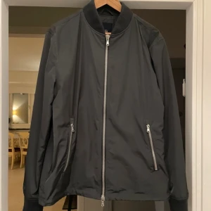 Mörkgrå  bomberjacka från Riley - Snygg mörkgrå bomberjacka från Riley med silvriga dragkedjor framtill och på fickorna. Klassisk ribbad krage och muddar vid ärmslut och nederkant. Tillverkad i ett slätt, lätt material som ger en clean och modern look.