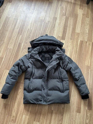 Canada goose  - Snygg mörkgrå dunjacka från Canada Goose med avtagbar huva och klassisk patch på ärmen. Jackan har dragkedja och knappar framtill samt två stora fickor. Perfekt för kalla vinterdagar och riktigt varm