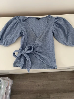 Blå omlottopp med puffärm och knyt - Supersöt ljusblå omlottopp med puffiga trekvartsärmar och knyt i midjan. Toppen har en v-ringad hals och ett strukturerat tyg som ger en unik look. Perfekt för dig som vill ha något trendigt och bekvämt.