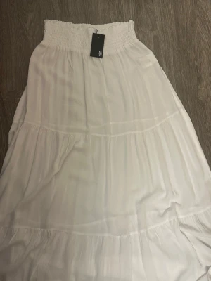 Vit långkjol med smock från LS - Luftig vit långkjol från Lager 157 med bred smock i midjan och volangdetaljer. Kjolen har en flowy passform och är perfekt för varma dagar. Tillverkad i ett mjukt och lätt material som ger en avslappnad känsla. Lång(PRISET GÅR ATT DISKUTERA)