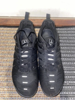 Nike Air Vapormax Plus svarta sneakers - Säljer ett par svarta Nike Air Vapormax Plus sneakers med reflekterande detaljer på ovandelen och genomskinlig sula. Skorna har en sportig design med plastdetaljer på sidorna och klassisk Nike-logga på plösen. Perfekta för dig som gillar streetwear och snygga detaljer. Skick som på bilderna, har ett hål längst fram på ena skorn vid tårna och hål under skorna.