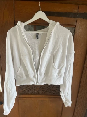 Vit cropped hoodie från Divided - Säljer en vit cropped hoodie från H&M Divided i storlek L. Hoodien har dragkedja framtill, två fickor och ribbade muddar. Tillverkad i mjukt sweatshirtmaterial och har en avslappnad passform med huva. Känns som S men står L