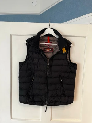 Svart dunväst från Parajumpers - Snygg svart dunväst från Parajumpers med dragkedja framtill och två fickor med dragkedja. Västen har en hög krage med gul detalj och klassisk Parajumpers-logga på nacken. Perfekt för lager-på-lager och riktigt skön att bära. En liten skada, se bild 3