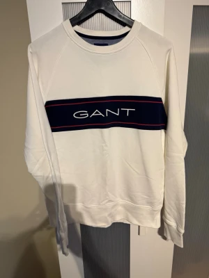 Vit sweatshirt från GANT - Säljer en stilren vit sweatshirt från GANT med marinblått band och röd detalj över bröstet där loggan är broderad. Tröjan har rund halsringning och ribbade muddar vid ärmslut och nederkant. Perfekt för en clean och sportig look.