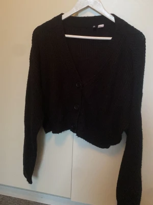 Svart cropped kofta med knappar - Säljer en svart, stickad cropped kofta med v-ringning och tre knappar framtill. Koftan har långa ärmar och en avslappnad passform, perfekt att slänga över en t-shirt eller linne. Materialet känns mjukt och mysigt.