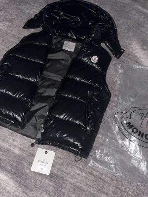 Svart dunväst från Moncler - Snygg svart dunväst från Moncler med glansig finish och avtagbar huva. Västen har två dragkedjeförsedda fickor och klassisk Moncler-logga på bröstet. Perfekt för lager-på-lager och streetwear-stil. Materialet är dunfyllning och yttertyget är blankt.