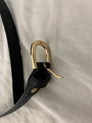 Svart bälte med guldfärgat spänne - Stilrent svart bälte i skinn med ett unikt guldfärgat metallspänne. Perfekt accessoar för att lyfta din outfit med en elegant touch. Bältet har klassisk design och smal rem som passar till både jeans och byxor.