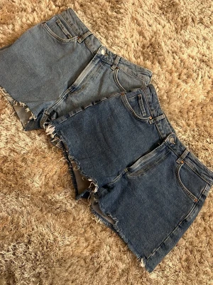 Blåa jeansshorts med rå kant - Säljer två par klassiska blå jeansshorts med rå, fransig kant och femficksmodell. Shortsen har hög midja och stängs med knapp och dragkedja. Perfekta till sommaren och gjorda i slitstark bomull. Båda shortsen är i storlek S och är från Bik Bok. Ena shortsen är lite ljusare och de andra lite mörkare.