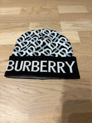 Svartvit Burberry mössa med logga - Snygg stickad mössa från Burberry i svart och vitt med stor logga framtill. Mössan har ett grafiskt mönster i vitt och ljusblått över hela ytan och är tillverkad i mjukt material som håller dig varm. Perfekt accessoar för att lyfta din outfit.