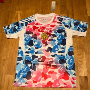 Adidas x BAPE camo special jersey - Säljer en Adidas x BAPE t-shirt med färgglatt camomönster i blått, rött, rosa och vitt. T-shirten har korta ärmar, klassiska Adidas-ränder på axlarna och är gjord i polyester. Perfekt för dig som gillar streetwear och unika samarbeten.