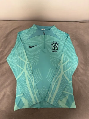 Nike Brasil träningströja grön - Snygg turkos långärmad träningströja från Nike med Brasil-logga och CBF-märke på bröstet. Tröjan har halv dragkedja framtill och ett grafiskt mönster i ljusblått och vitt. Perfekt för fotbollsträning eller sportiga aktiviteter.