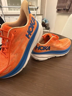 Hoka Clifton 9 orange sneakers - Snygga Hoka Clifton 9 sneakers i orange med blå detaljer och vit/blå sula. Ovandelen är i mesh för bra ventilation och komfort. Perfekta för dig som gillar sportig stil och vill ha riktigt lätta och bekväma skor. Tydlig Hoka-logga på sidan.