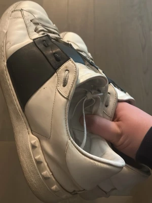 Valentino Open  - Snygga vita sneakers med bred svart läderdetalj över sidan. Klassisk låg modell med vita snören och coola nitar runt sulan. Perfekta för dig som gillar stilrena men ändå unika skor. Passar till det mesta och ger en clean look. 