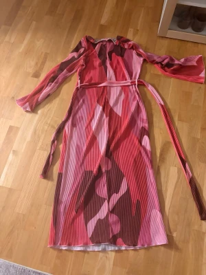 Mönstrad omlottklänning i rosa och rött - Säljer en långärmad klänning med bälte i midjan. Klänningen har ett färgstarkt abstrakt mönster i rosa, rött och brunt, samt plisserat tyg som ger en cool struktur. Perfekt för dig som gillar statement-plagg och vill sticka ut.