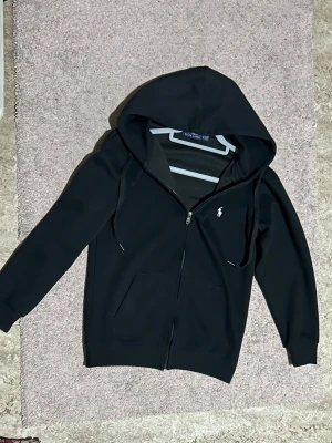 Svart hoodie från Ralph Lauren - Svart hoodie från Ralph Lauren med dragkedja framtill och klassisk vit broderad logga på bröstet. Hoodien har huva med dragsko och två fickor framtill. Tillverkad i mjukt bomullsmaterial som känns skönt mot huden. Perfekt för en avslappnad och stilren look.