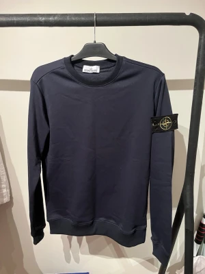 Mörkblå sweatshirt från Stone Island - Snygg mörkblå sweatshirt från Stone Island med klassisk rund hals och ribbade muddar. Ikonisk logopatch på vänster ärm. Perfekt för dig som gillar stilrena och exklusiva plagg. Materialet är mjukt och bekvämt, passar till många olika outfits.