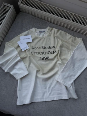 Vit långärmad topp Acne Studios - Vit långärmad topp från Acne Studios med tryck 'Acne Studios STOCKHOLM 1996' på bröstet. Klassisk rund halsringning och avslappnad passform. Tillverkad i mjuk bomull, perfekt för en clean och stilren look.