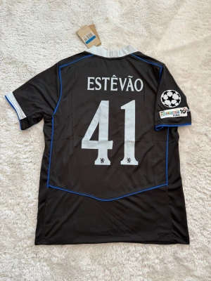 Chelsea svart matchtröja Nike - Säljer en svart Chelsea matchtröja från Nike med Estêvão och nummer 41 på ryggen. Tröjan har blå detaljer, grå krage och ärmar samt Champions League och FIFA-märken på ärmen och bröstet. Perfekt för dig som älskar fotboll och vill sticka ut på planen.