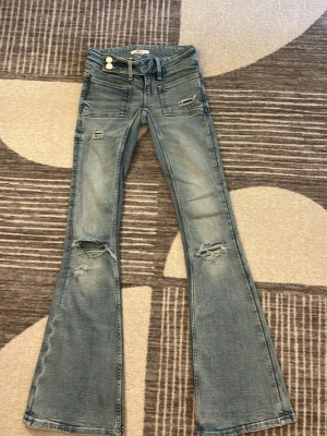 Blå bootcut jeans med slitningar - Säljer ett par ljusblå bootcut jeans med slitna detaljer och råa hål på benen. Jeansen har låg midja, fem fickor och dubbla knappar fram. Materialet är klassisk denim i bomull. Perfekta för en avslappnad och trendig look.