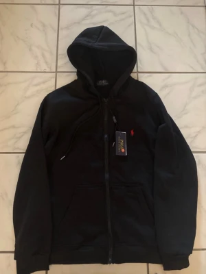 Svart hoodie från Polo Ralph Lauren - Snygg svart hoodie från Polo Ralph Lauren med dragkedja framtill och klassisk röd logga broderad på bröstet. Hoodien har huva med dragsko och kängurufickor. Tillverkad i mjukt material som känns skönt mot huden. Perfekt för en avslappnad och stilren look. Priset kan diskuteras i dm. 