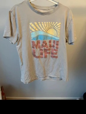Grå Maui Life t-shirt med tryck - Snygg beige t-shirt med färgglatt Maui Life-tryck framtill som visar sol, hav och text. Klassisk rund hals och korta ärmar. Perfekt för dig som gillar avslappnad stil och vill ha en unik t-shirt med somriga vibbar.