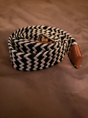 Blått vitt flätat bälte - Snyggt flätat bälte i svart och vitt med bruna läderdetaljer vid ändarna. Bältet har ett randigt mönster. Passar till jeans eller chinos och är lätt att justera efter önskad längd.