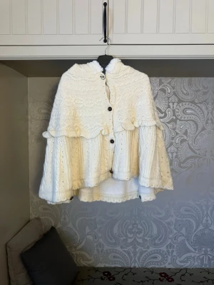 Vit stickad poncho med huva Axes femme - Supersöt vit stickad poncho från axes femme med fluffigt foder och stor huva. Ponchon har dekorativa knappar med rosett, volangkant och kabelstickat mönster. Aldrig använd.