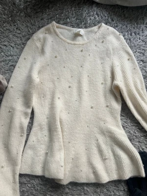 Vit stickad tröja med pärlor - Säljer en vit stickad tröja med rund hals och långa ärmar. Tröjan har ett ribbat mönster och är dekorerad med små pärlor över hela plagget, vilket ger en elegant touch. Perfekt för dig som gillar stilrena detaljer och mjuka material.