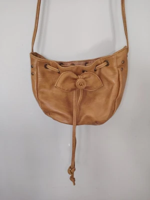 Beige axelväska med rosett från Blanco - Söt beige axelväska från Blanco Accessories i mjukt konstläder. Väskan har en dekorativ rosett framtill, dragskostängning med snören och metallöljetter. Invändigt finns ett fack med dragkedja. Perfekt för dig som gillar en feminin och enkel stil. Är i fint använt skick, några skavanker som syns på bild. 