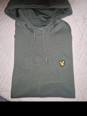 Olivgrön hoodie från Lyle & Scott - Snygg olivgrön hoodie från Lyle & Scott med klassisk gul logga på bröstet. Hoodien har dragsko i huvan och är tillverkad i mjukt material som känns skönt mot huden. Perfekt för en avslappnad och stilren look.