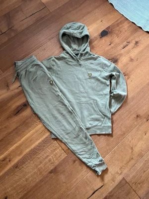 Grönt loungewear-set med luva - Snyggt loungewear-set i ljusgrönt från Lyle & Scott. Setet består av en hoodie med luva och känguruficka samt matchande byxor med dragsko och muddar. Båda plaggen har den klassiska gula loggan broderad på bröstet och benet. Perfekt för chill dagar hemma.