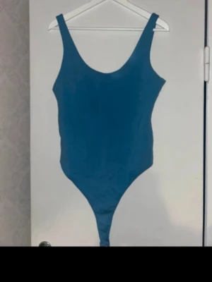 Blå body med breda axelband - Säljer en stilren blå body med breda axelband och rundad urringning. Modellen är enkel och tight, vilket ger en clean look. Materialet ser stretchigt och mjukt ut, perfekt för att sitta skönt mot kroppen. Inga synliga detaljer eller märke.