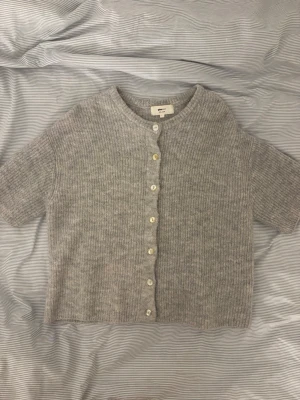 Grå stickad kofta från Gina Tricot - Kort, grå stickad kofta från Gina Tricot i ullblandning. Koftan har rund halsringning, korta ärmar och vita knappar framtill. Ribbstickad struktur ger en mysig och trendig look. Perfekt att slänga över en topp för extra lager.