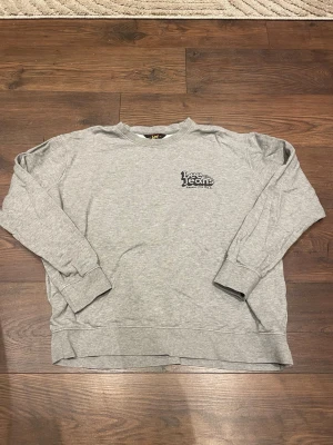 Grå sweatshirt från Lee Jeans - Säljer en klassisk grå sweatshirt från Lee Jeans med tryckt logga på bröstet. Tröjan har rund halsringning, långa ärmar och ribbade muddar vid ärmslut och nederkant. Perfekt för en avslappnad och stilren look.