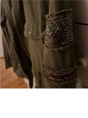 Olivgrön utilityjacka med pärldetaljer - Snygg olivgrön utilityjacka från CIMINY med dekorativa pärlbroderier på ena ärmen. Jackan har flera fickor med knappar, snörning i midjan och axelklaffar. Perfekt för dig som vill ha en unik och cool look.