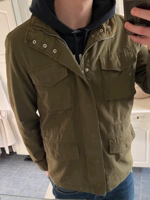 Grön fieldjacket - Säljer en olivgrön parkajacka med fyra stora fickor framtill och dragkedja. Jackan har en klassisk militärinspirerad look och är tillverkad i bomullsmaterial. Perfekt för dig som gillar streetwear och vill ha en stilren jacka till vardags.