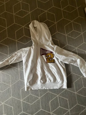 Vit hoodie från Dsquared2 STORLEK XS men sitter som S !!!!!!!!! - Säljer en vit hoodie från Dsquared2 med stor huva och magficka. På bröstet finns ett stort Dsquared2-tryck i vinrött och gult. Hoodien har långa ärmar och ribbade muddar. Perfekt för en avslappnad och sportig stil.