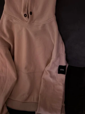Beige hoodie från Jack&jones - Säljer en beige hoodie från jack&jones med klassisk känguruficka och huva med metallöljetter. Tröjan har en svart patch med logga på ena ärmen och är gjord i mjukt bomullsmaterial. Perfekt för en chill och avslappnad stil.