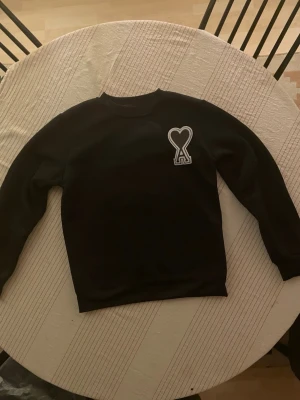 Svart sweatshirt från Ami Paris - Säljer en svart sweatshirt från Ami Paris med det ikoniska hjärta-A-märket broderat på bröstet. Tröjan har rund hals, långa ärmar och är gjord i mjukt bomullsmaterial. Perfekt för dig som gillar stilrena och trendiga plagg med en twist. 