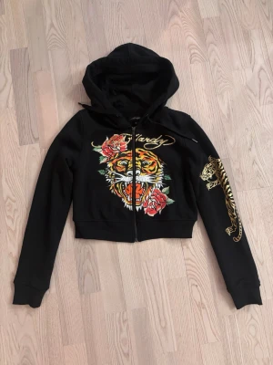 Svart Ed Hardy hoodie med tigertryck - Snygg svart hoodie från Ed Hardy med dragkedja, huva och croppad passform. Framsidan har ett stort tigertryck med röda rosor och guldtext, medan ena ärmen pryds av en guldtiger. Baksidan har Ed Hardy-logga i guld. Perfekt för dig som gillar streetwear och statement-plagg.