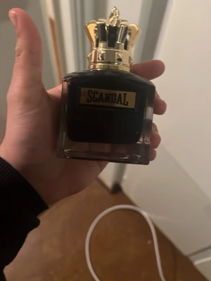 Scandal parfym med gulddetaljer - Lyxig parfymflaska från Scandal med mörk glasflaska och en unik guldfärgad krona som lock. Flaskan har en elegant och tung design med svart etikett och guldtext. Perfekt för dig som gillar exklusiva dofter och snygg inredningsdetalj på hyllan.upphämtning endast